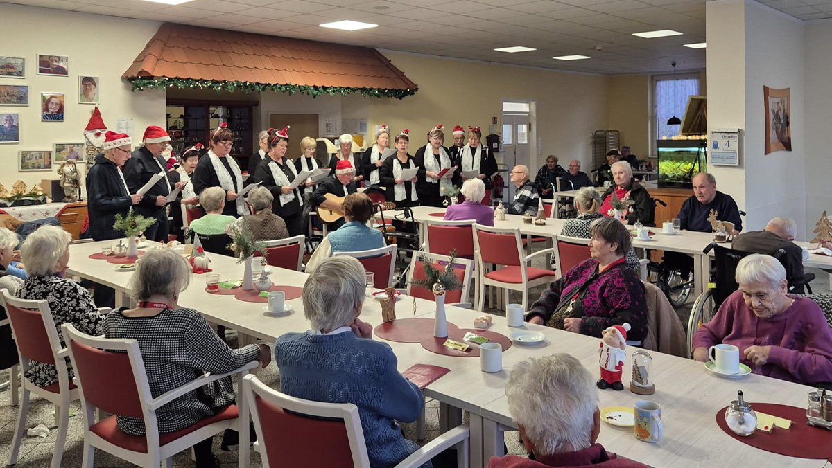 Der Damsdorfer Chor tritt im Gemeinschaftsraum der ProCurand Seniorenresidenz Am G&ouml;rnsee auf, w&auml;hrend die Bewohner*innen an den Tischen zuh&ouml;ren.