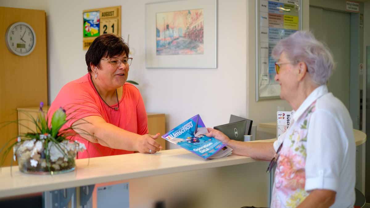 Die Rezeptionistin im Service-Wohnen der Seniorenresidenz Am Hufeisensee empfängt am Tresen eine Seniorin.
